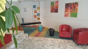 Een hal met een marmeren vloer, een zwarte tafel met stoelen, een rode stoel, een prullenbak en kleurrijke kunstwerken aan de muren.