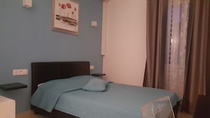 Een slaapkamer met een bed, nachtkastje, een lichtschakelaar aan de muur, en een raam met gordijnen.