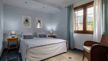 A bedroom with a bed, two bedside tables with lamps, a chair, a window with curtains, and artwork on the walls.