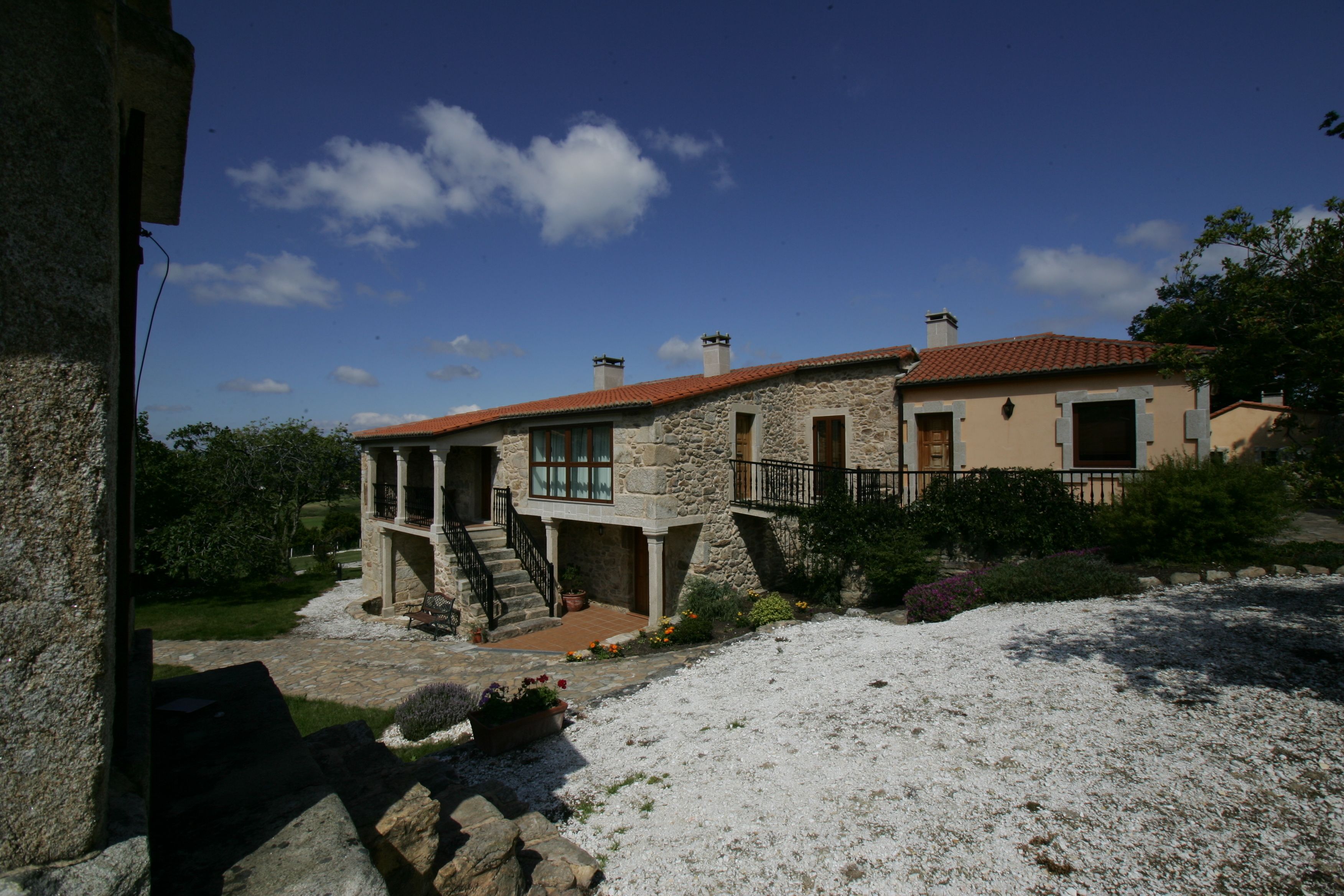 A stone house with a red-tiled roof, a stone staircase, and a garden with gravel and plants.