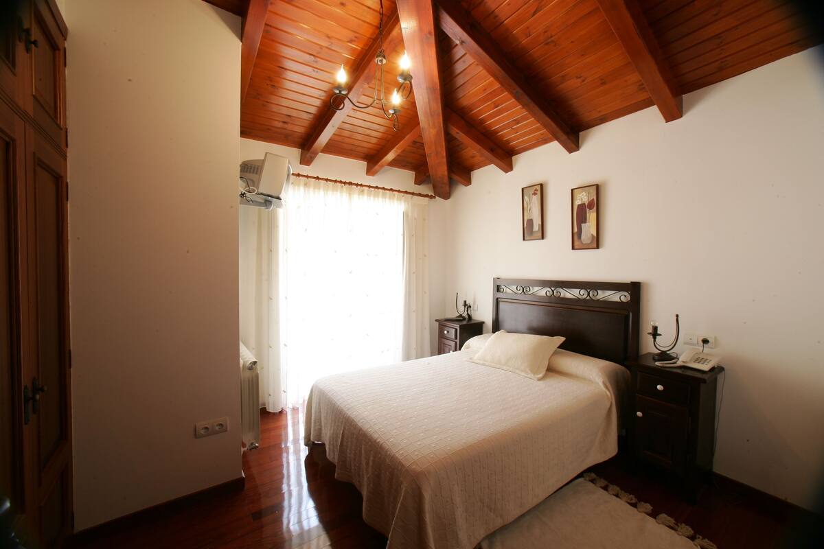 A bedroom with a wooden ceiling, a bed with a headboard, a nightstand, and a window with curtains.