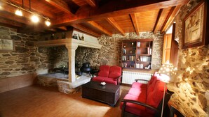 A cozy living room with stone walls, wooden beams, a fireplace, red upholstered chairs, a coffee table, and a bookshelf. - Casa da Botica (A Estrada)