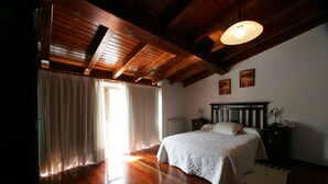 A bedroom with a wooden ceiling, a bed with a white bedspread, a nightstand, and a window with curtains. - Casa da Botica (A Estrada)