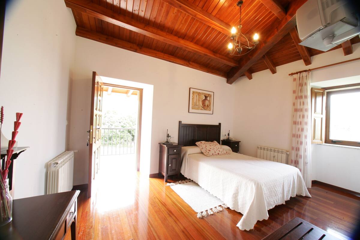 A bedroom with a wooden ceiling, a bed with white bedding, a nightstand, a radiator, and a window with curtains.