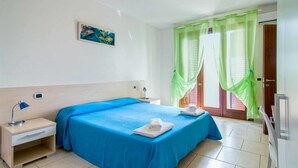 Une chambre d’hôtel avec un lit, deux tables de chevet, une petite table, une horloge numérique et un tableau au mur.