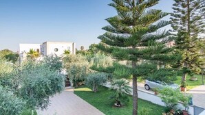 Un bâtiment moderne à la façade blanche, entouré d’un jardin bien entretenu où l’on trouve divers arbustes et un grand pin.
