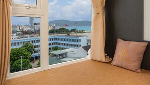 A room with a bed, beige curtains, a view of a cityscape, and a pillow with a logo. - Phuong Dong Hotel & Apartment (Quy Nhon Nam)