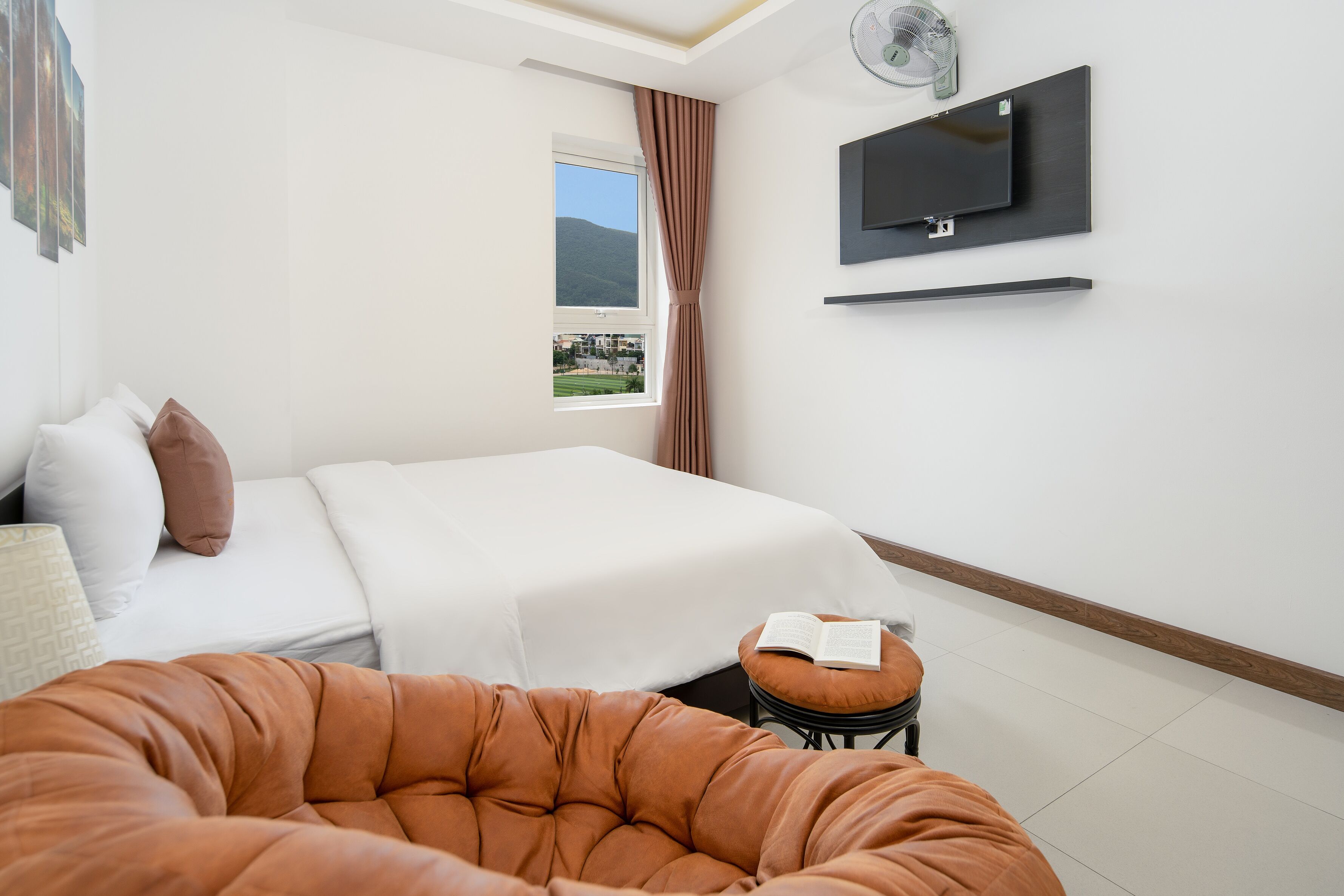 A modern hotel room with a bed, a reading chair, a TV mounted on the wall, and a window with a view of the outdoors.