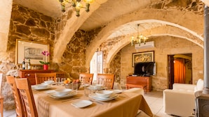 A stone-walled dining area with a table set for four, wooden chairs, a white sofa, and a television in the background. - TA Filomena (Xaghra)