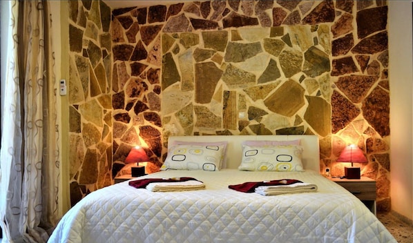 A bed with a white quilted bedspread, two bedside lamps, and a stone wall behind.