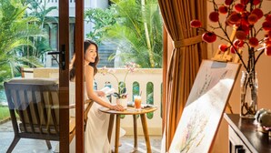 A woman seated at a table with a potted plant and a drink, overlooking a balcony with a view of greenery and a building. - Én Retreat Hoi An (Da Nang)