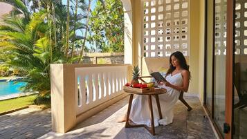 A woman sitting on a balcony with a view of a swimming pool and tropical plants. She is holding a book and there is a small table with fruits and a pineapple.