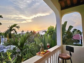 Un balcón con vista a un atardecer, una caja de madera con frutas y una copa de vino tinto, y una silla.