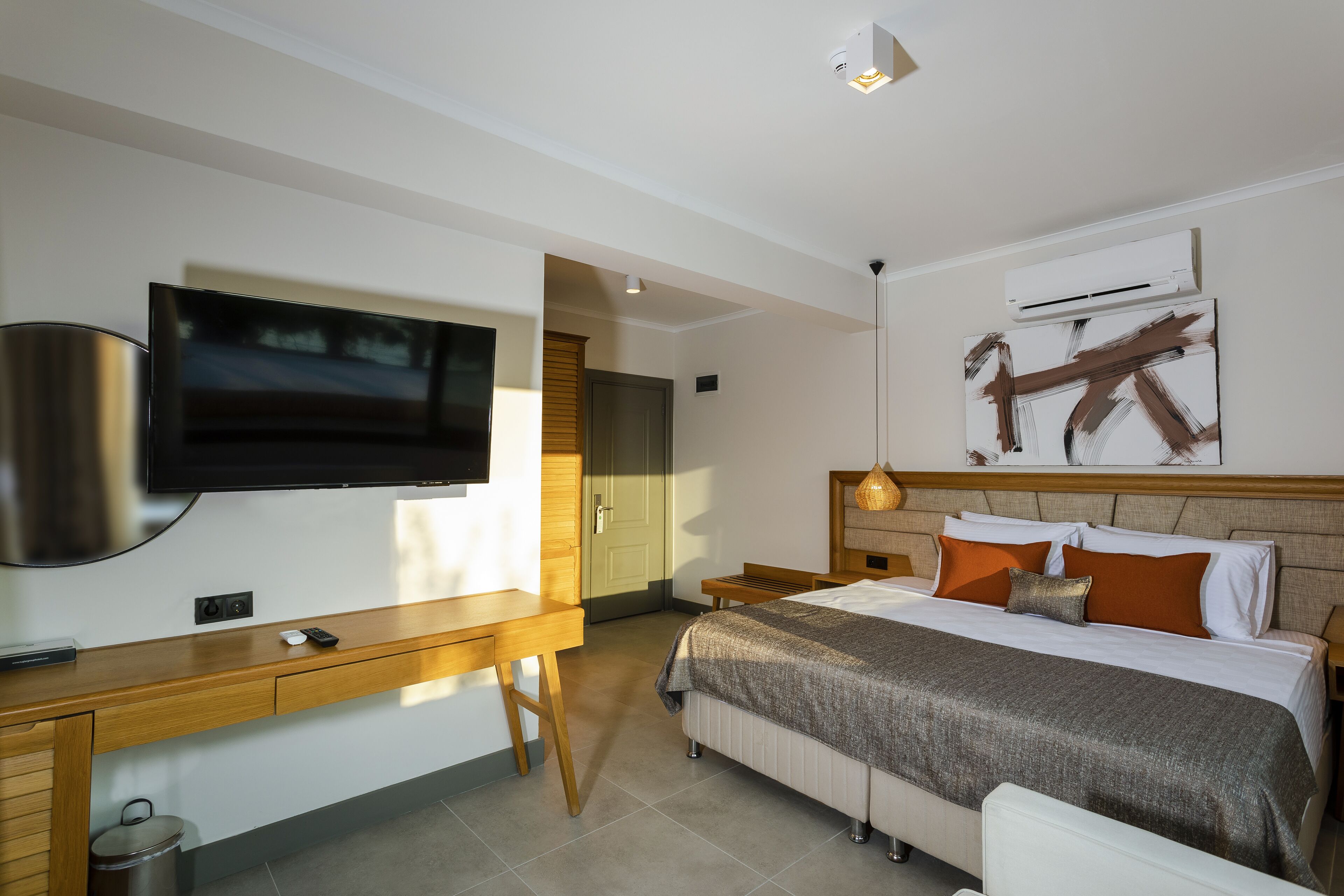 A modern hotel room with a flat-screen TV, wooden desk, bed with a grey and white bedspread, and a large abstract painting on the wall.