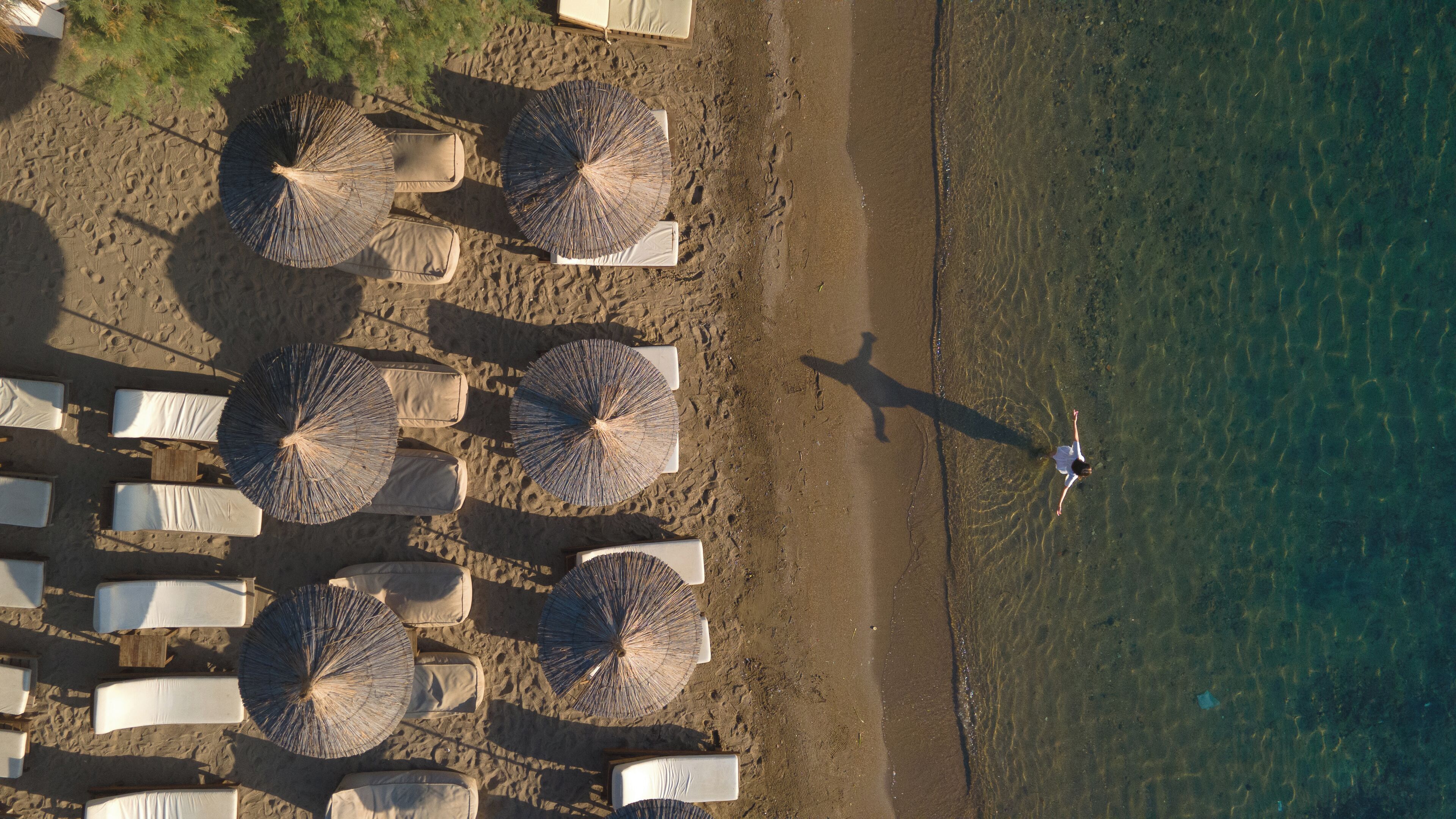 An aerial view of a beach with rows of umbrellas and lounge chairs, a person standing by the water, and clear shallow water.
