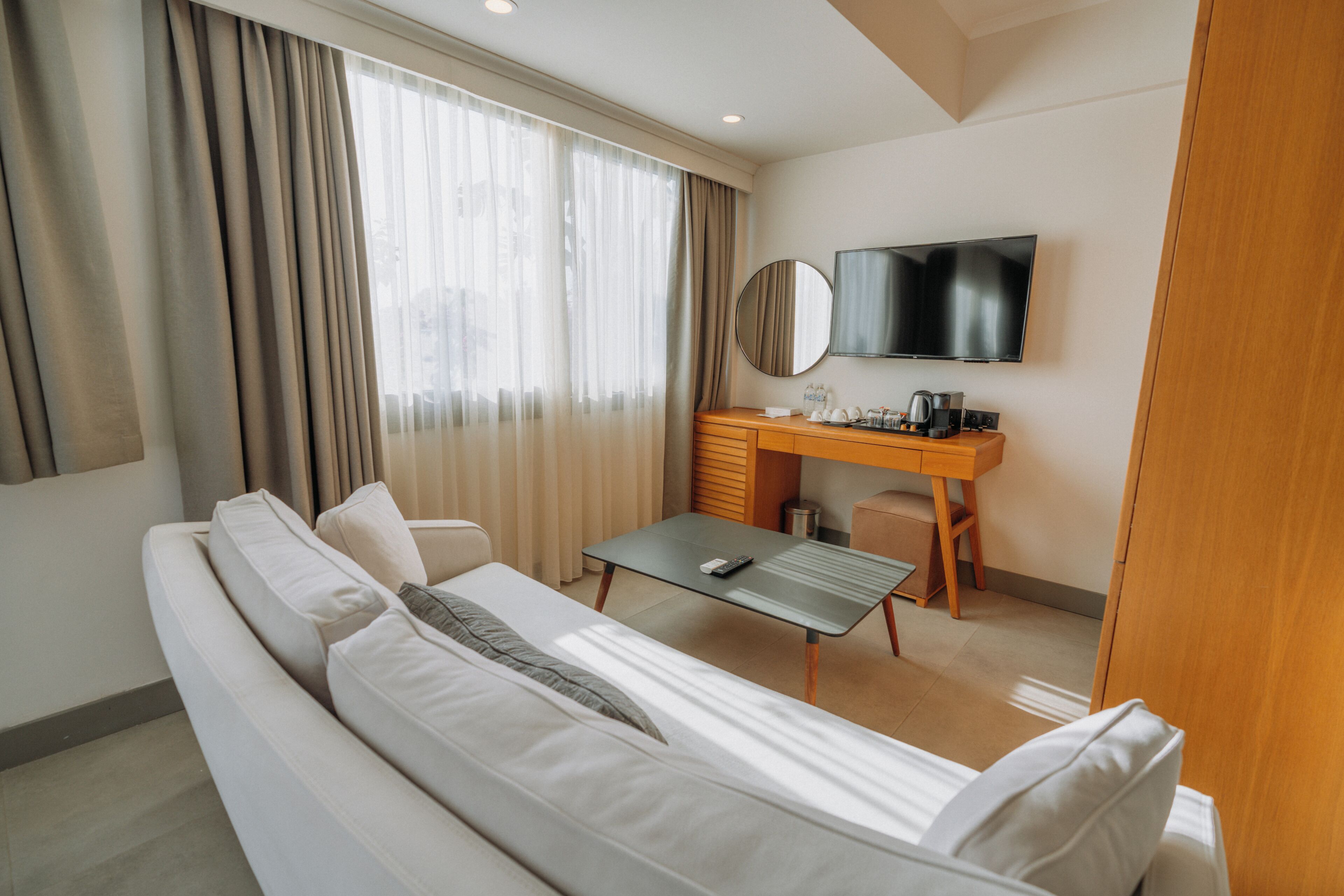 A modern hotel room with a sofa, a small table, a desk, a television, and a window with curtains.