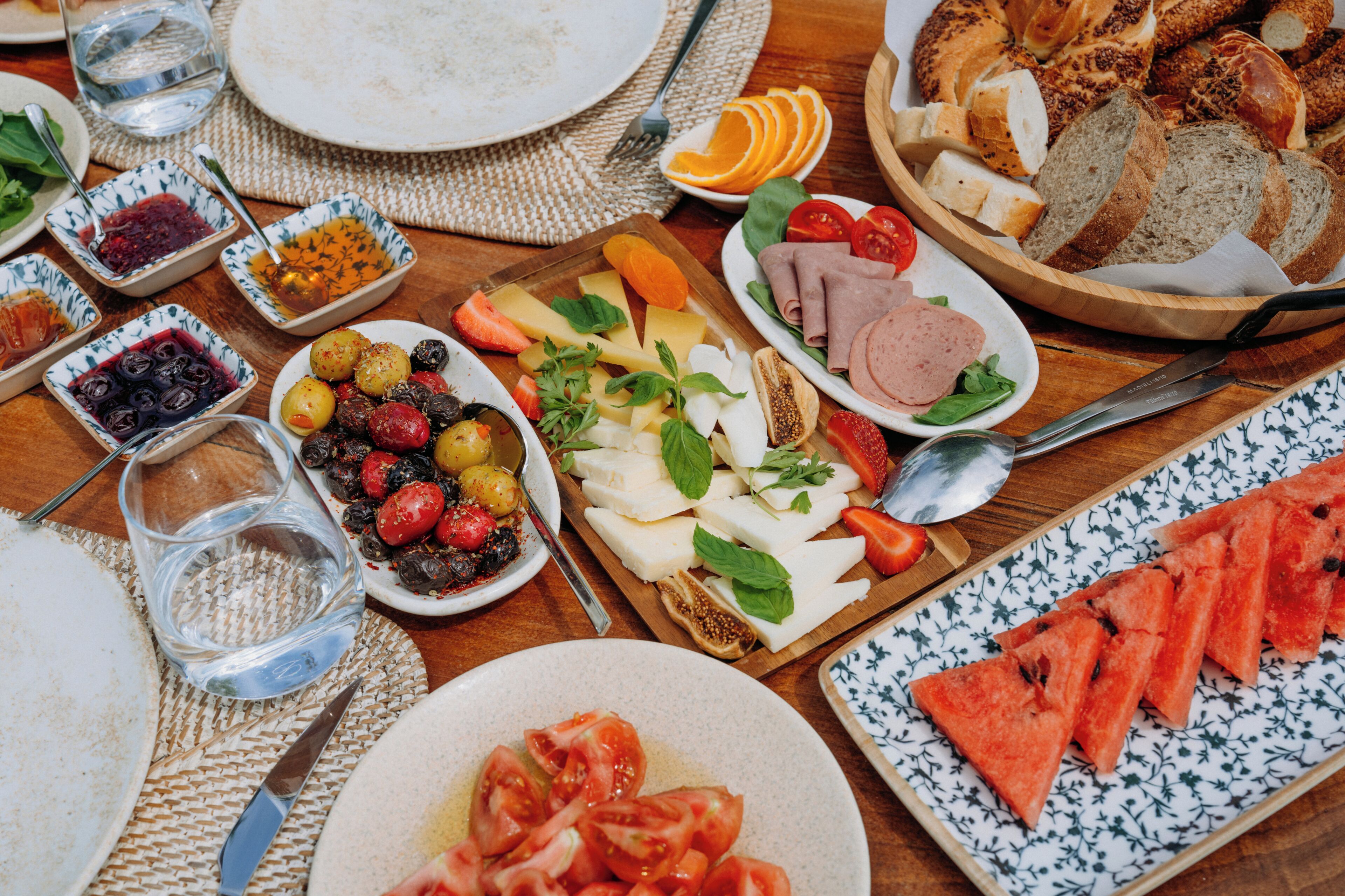 Een tafel met diverse etenswaren, zoals brood, kaas, tomaten en stukken watermeloen.