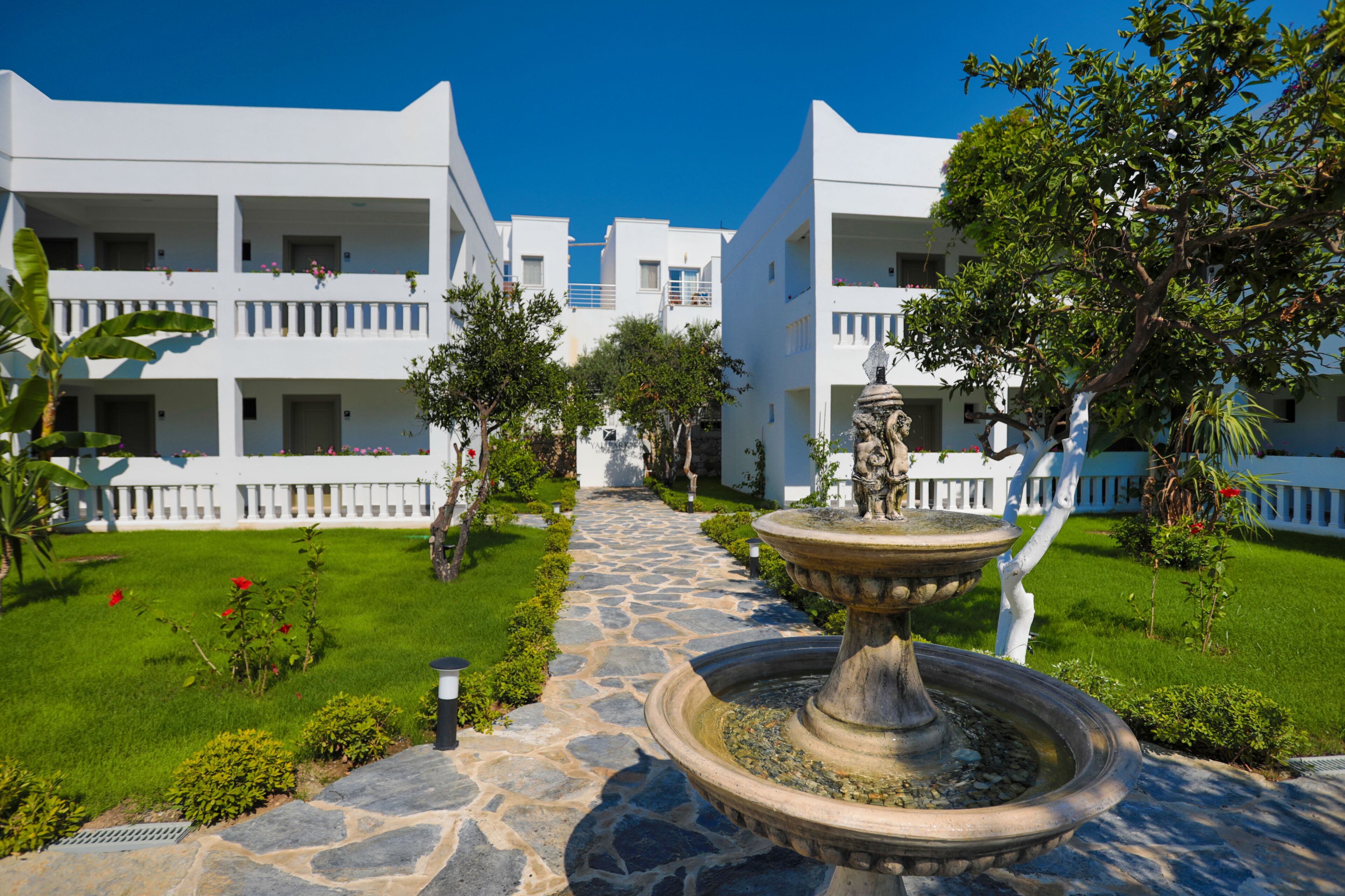 A courtyard with a stone pathway leading to a central fountain, surrounded by white buildings with balconies and lush greenery.