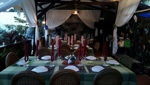 An outdoor dining area with tables set for a meal, surrounded by wicker chairs and draped with white fabric.