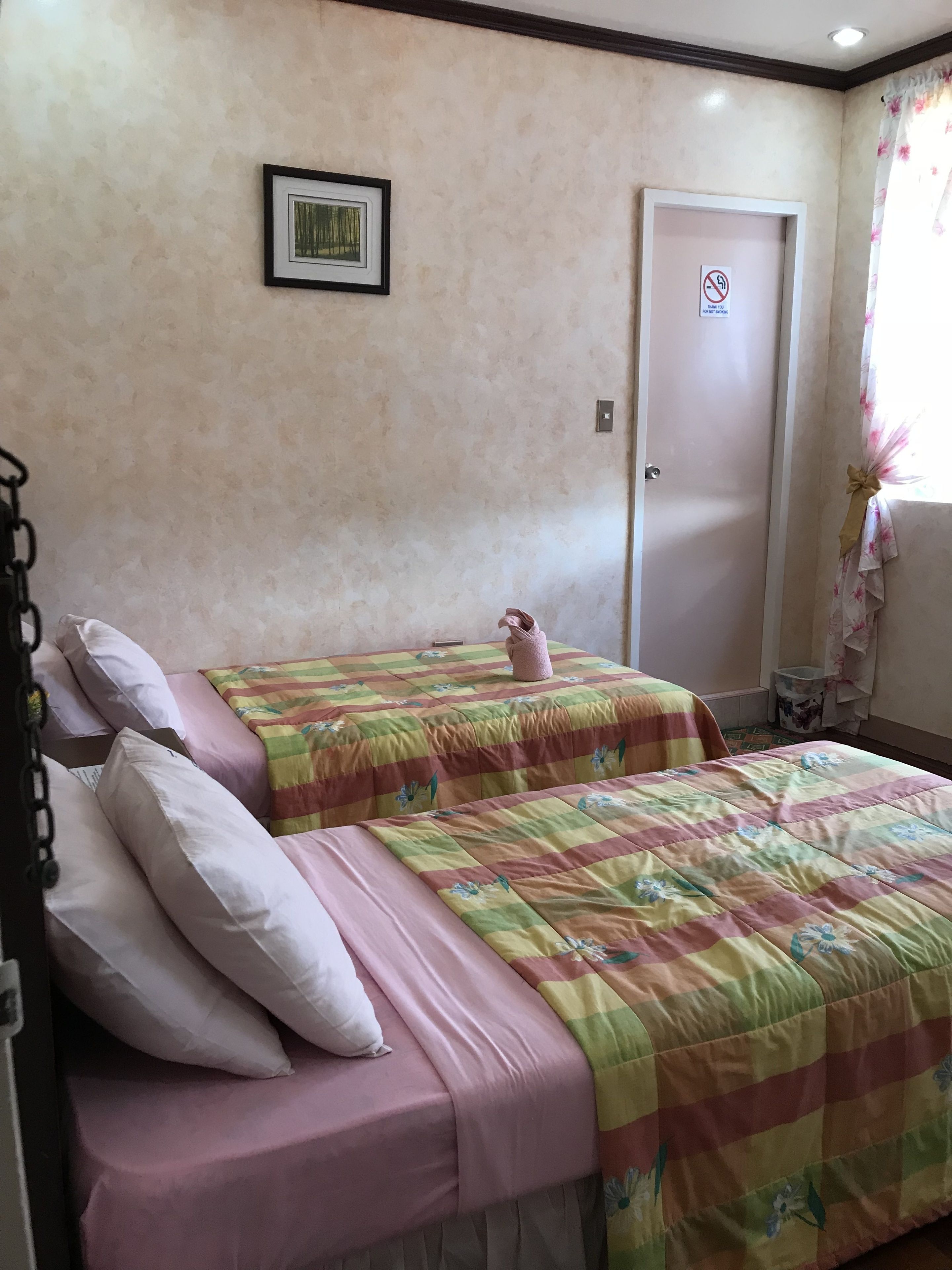 A double bed with pink and green floral bedding, a framed picture on the wall, and a door with a no-smoking sign.