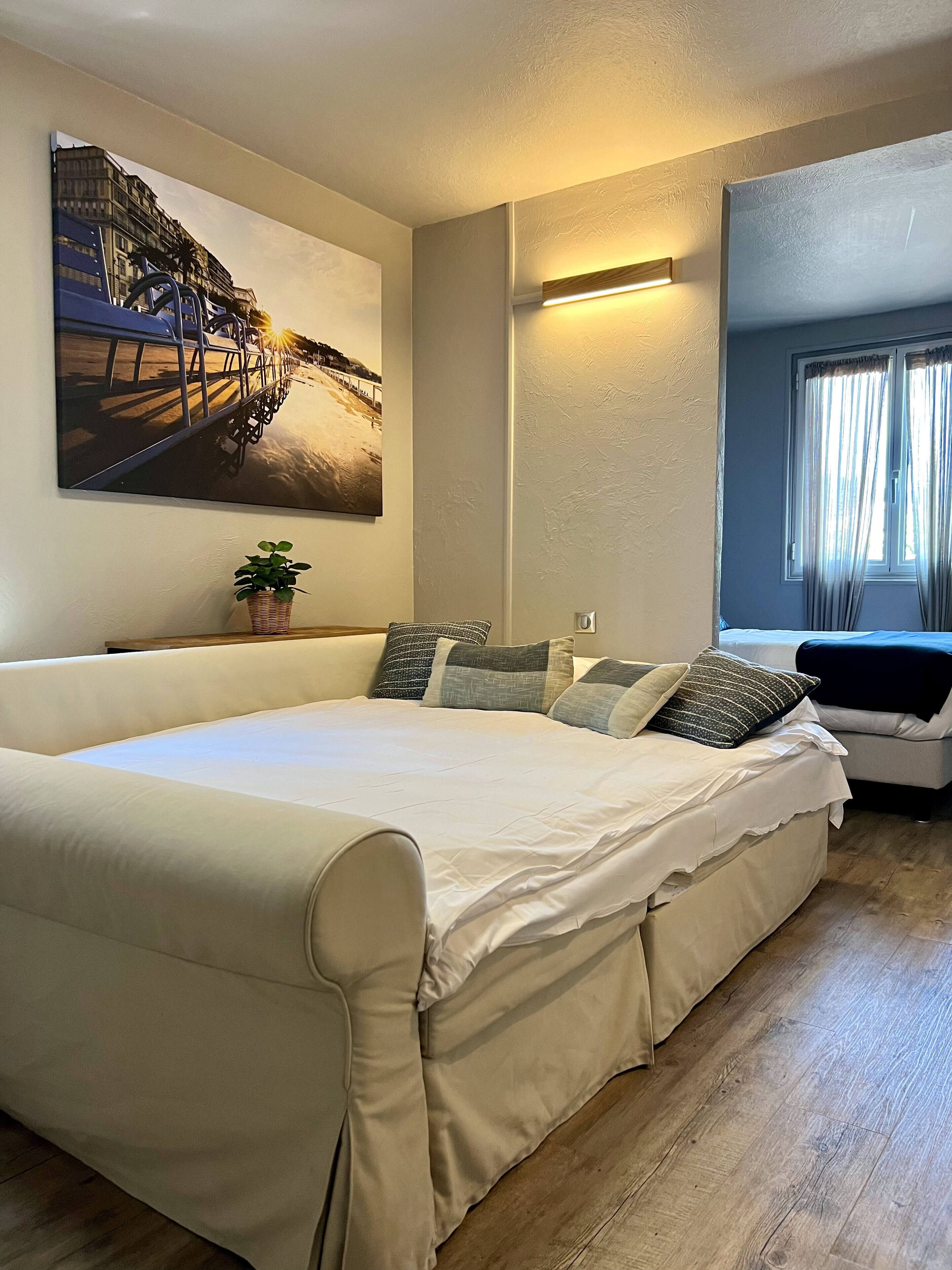 A bedroom with a large bed, a wall-mounted painting, a bedside table with a plant, and a window with curtains.
