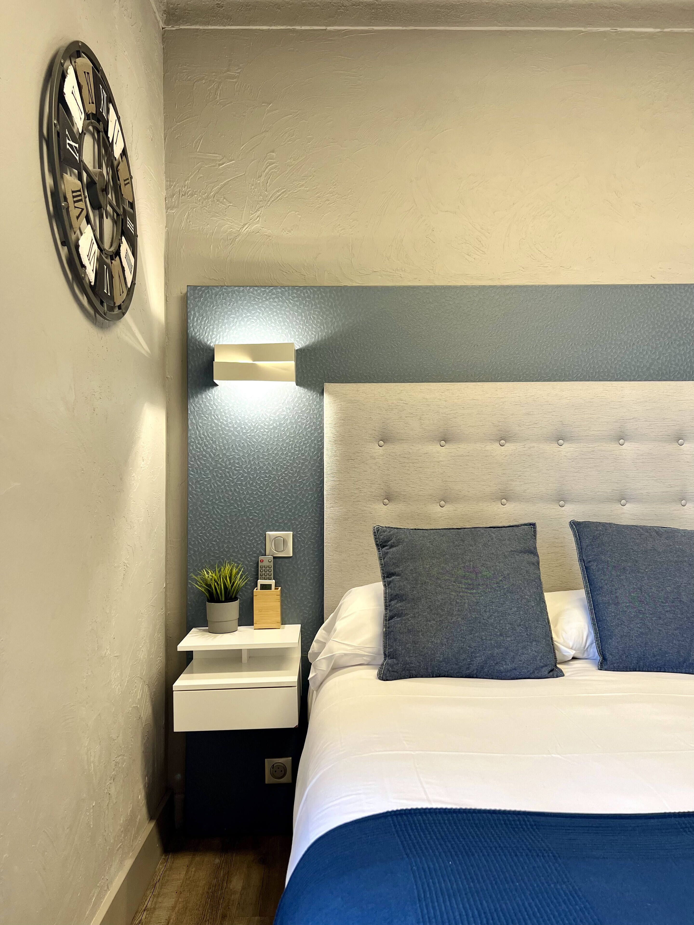 Un dormitorio con cama, mesita de noche, reloj de pared, planta pequeña e iluminación empotrada.