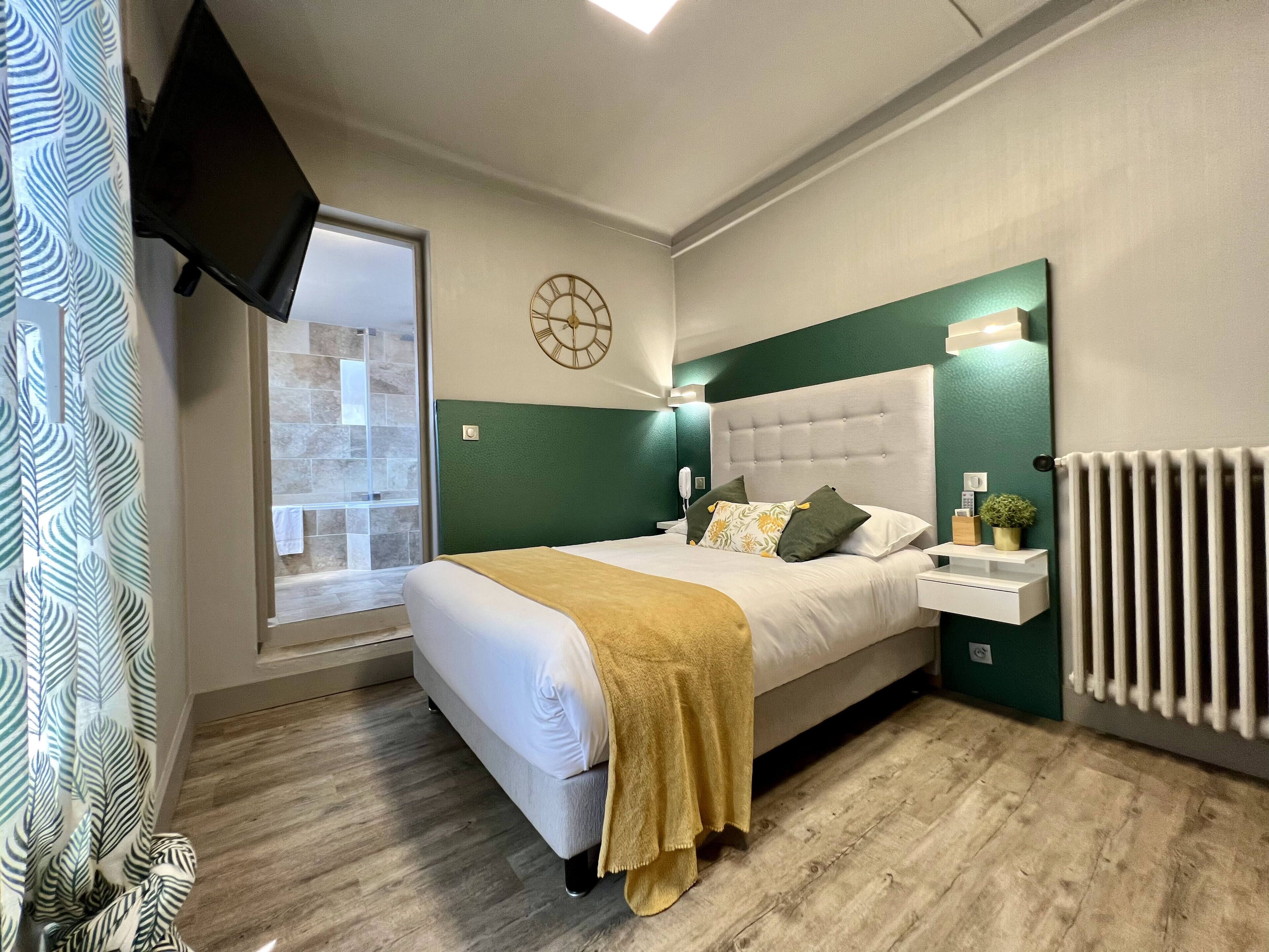 A modern bedroom with a large bed, a bedside table, a wall clock, and a window with a view of a stone wall.