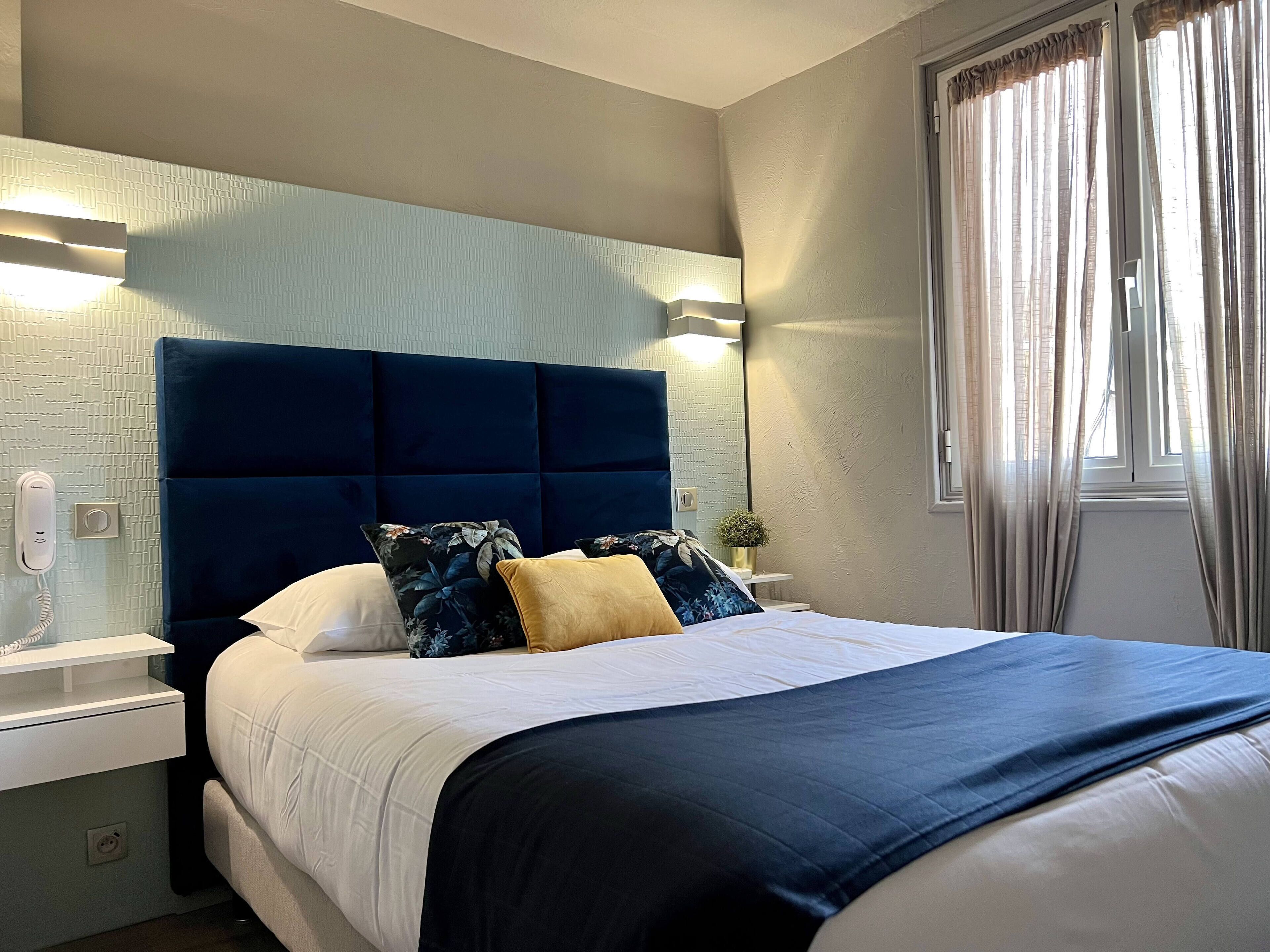 A neatly made bed with a dark headboard, white bedding, and decorative pillows. There is a bedside table with a lamp and a phone. A window with curtains is visible, and a small plant is on the nightstand.