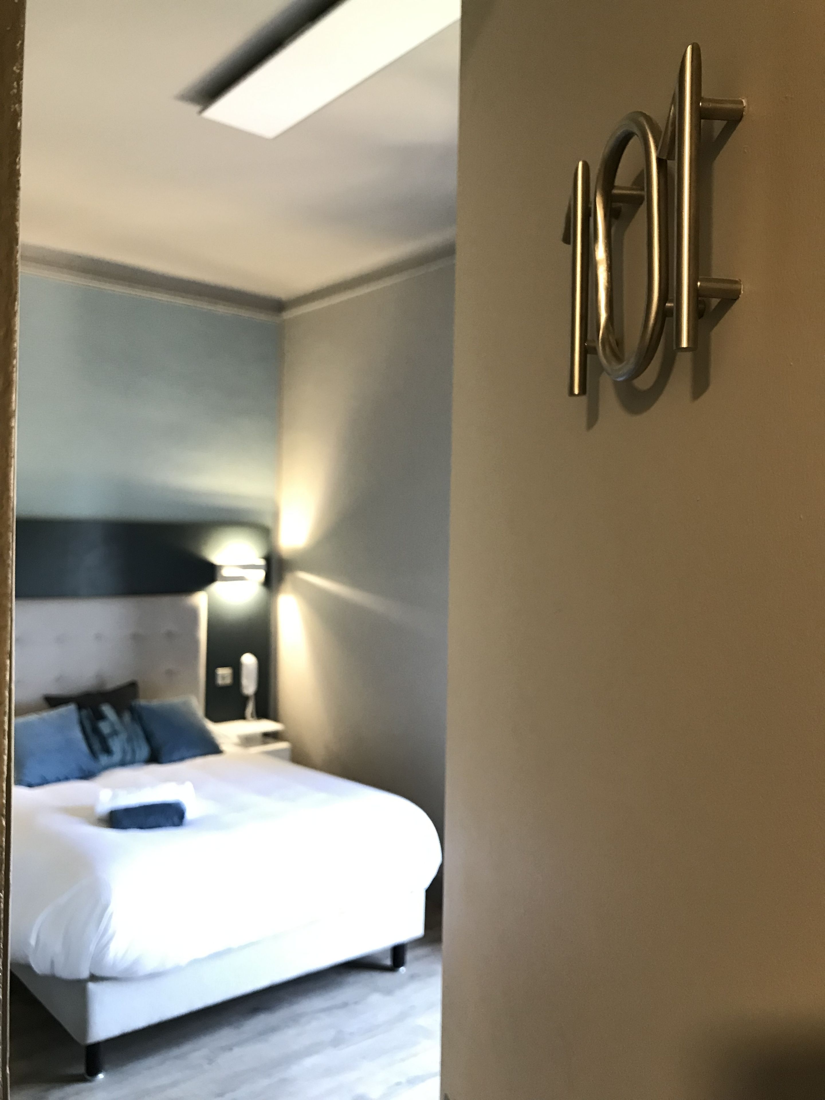 Ett hotellrum med en säng, ett nattduksbord, en lampa och en väggmonterad handduksställning.
