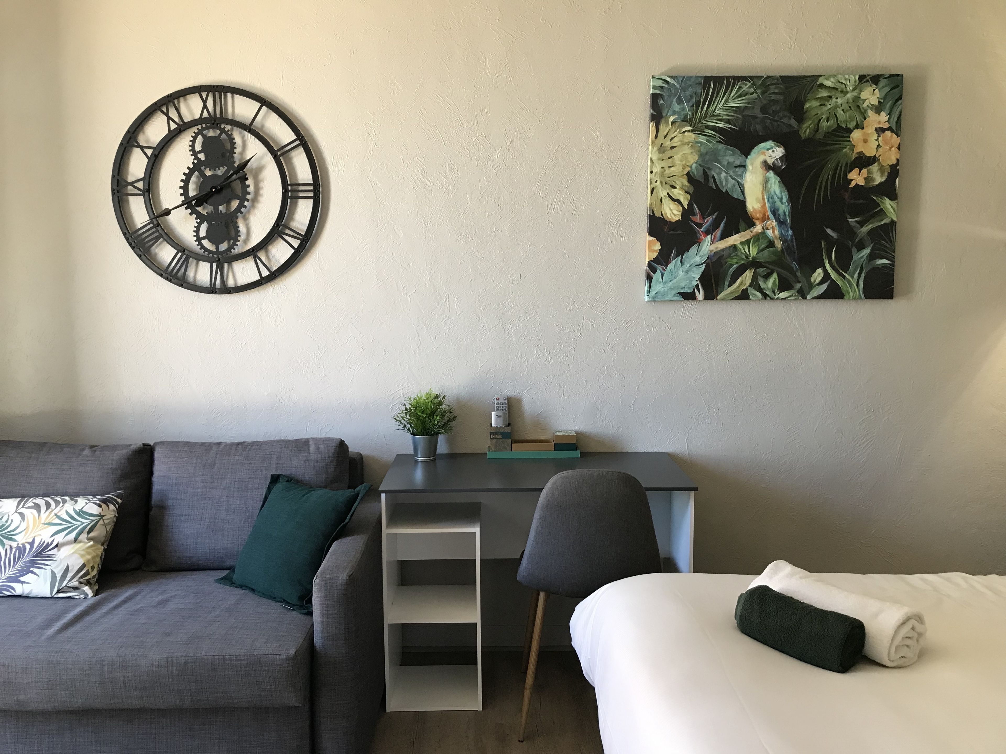 A modern room with a grey sofa, a desk with a plant, a wall clock, and a tropical painting.