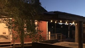 A wooden deck with a pergola, surrounded by greenery and a building with a red facade. - Chinova Mötesgård (Jarvso)