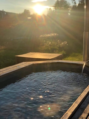 A wooden deck leading to a hot tub with water flowing into it, surrounded by greenery and a sunset in the background.