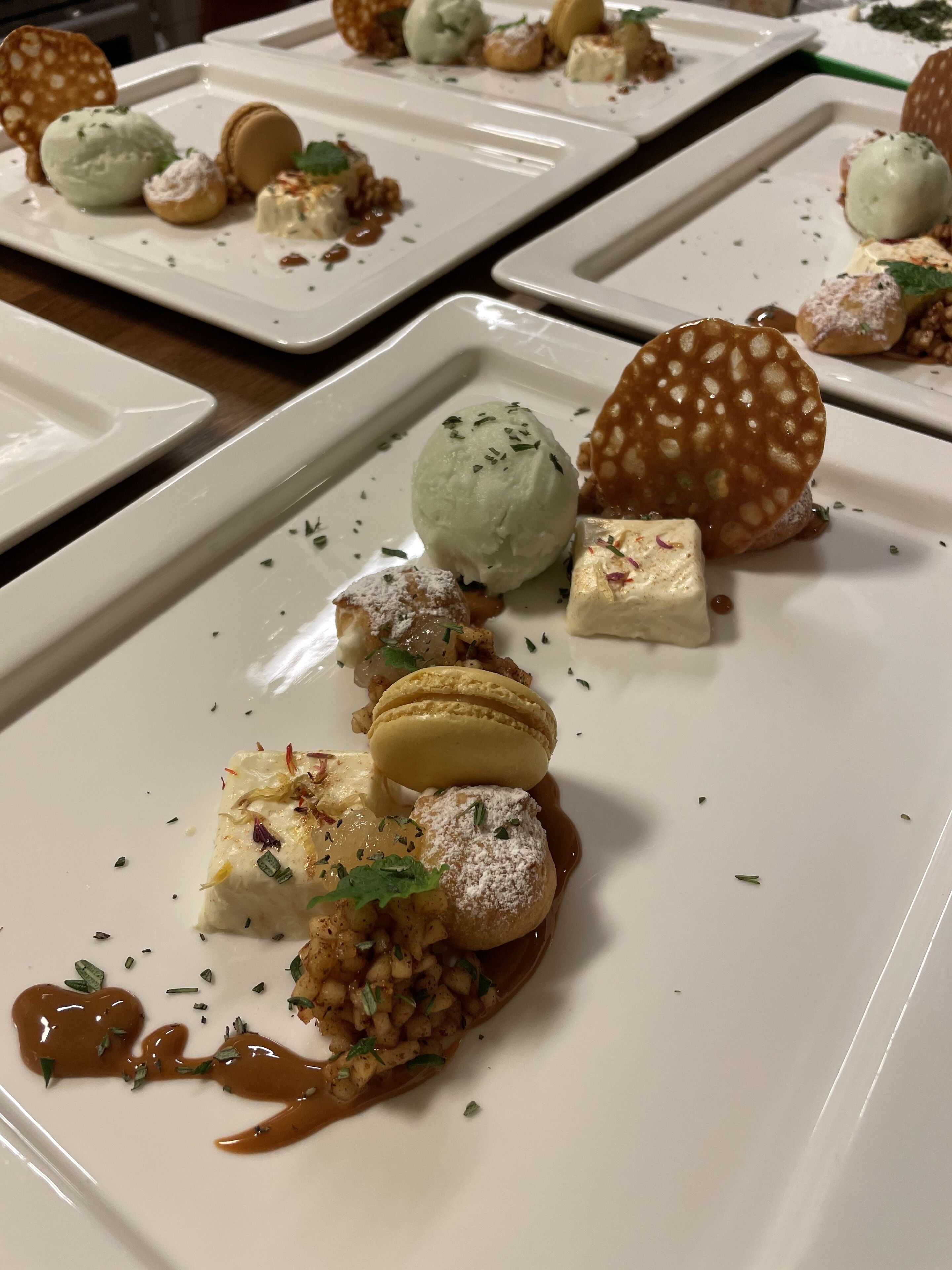 Une série d’assiettes carrées blanches, sur lesquelles sont présentés divers desserts, notamment des macarons, de la glace et d’autres gourmandises.