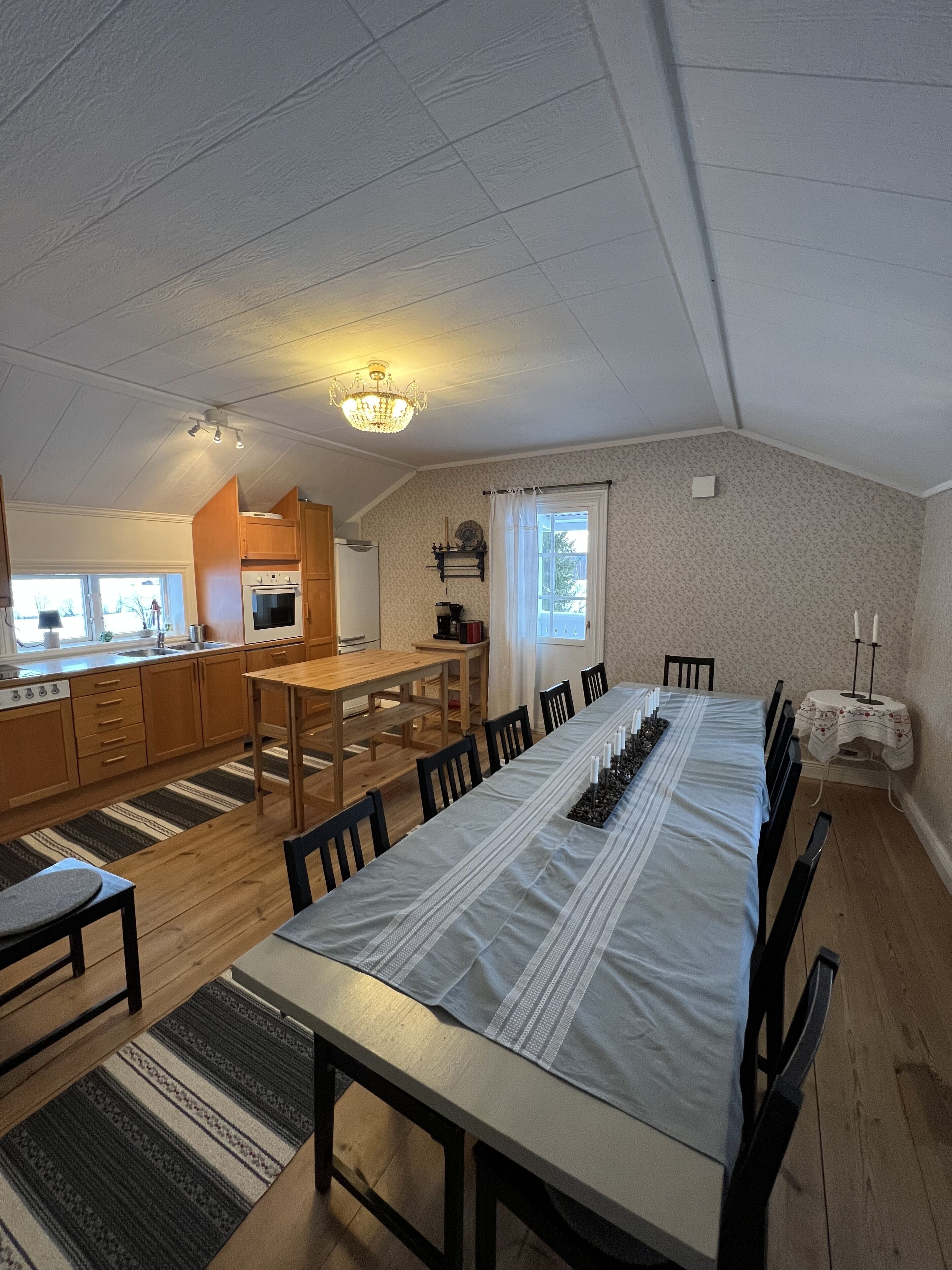 A spacious dining area with a long table set for six, wooden flooring, a chandelier, and a kitchen in the background.
