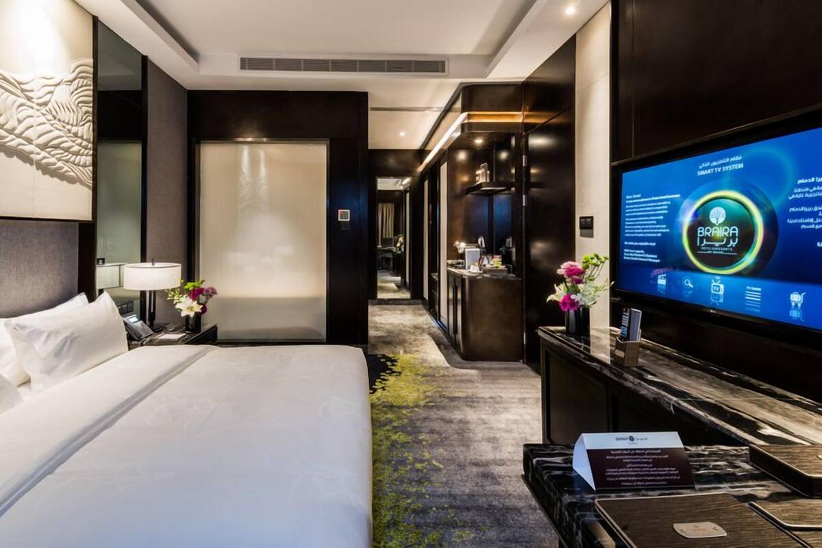 Une chambre d’hôtel avec un lit, un bureau doté d’un ordinateur, un téléviseur affichant le site web de l’hôtel, et un comptoir de réception en arrière-plan.