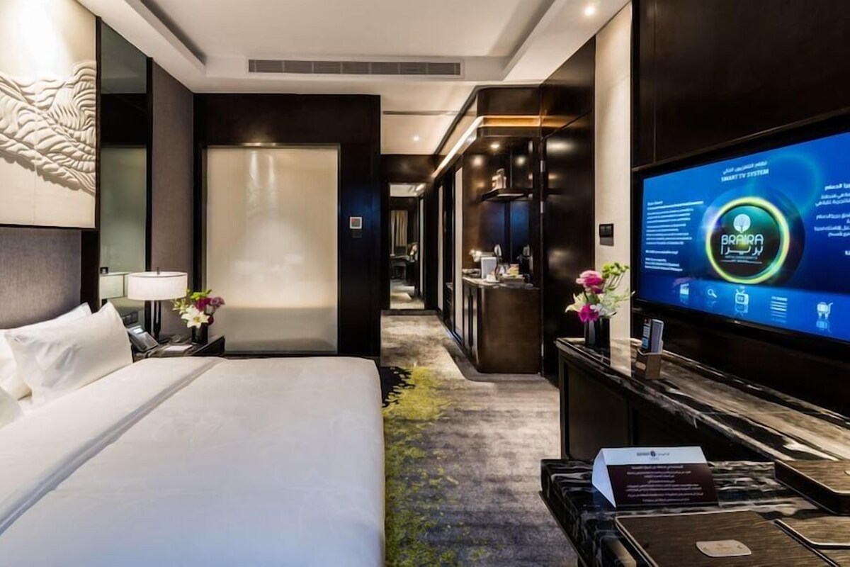 A modern hotel room with a large bed, a desk, a television displaying a digital interface, and a reception area in the background.
