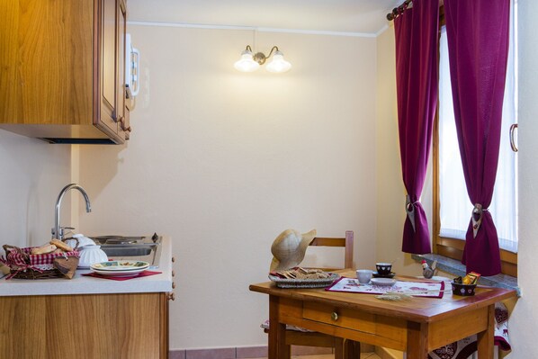 A kitchen with wooden cabinets, a sink, a window with purple curtains, and a dining table with a basket of bread and a kettle. - Agriturismo Lou Rosé (Donnas)