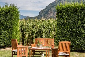 Une terrasse avec des chaises et une table en bois, entourée de grandes plantes vertes et avec une montagne en arrière-plan.