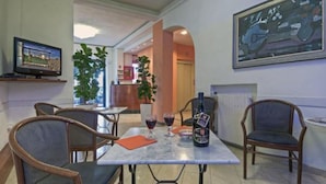 Un comedor de hotel con una mesa dispuesta para dos personas, dos sillas, una botella de vino y un televisor en un estante.