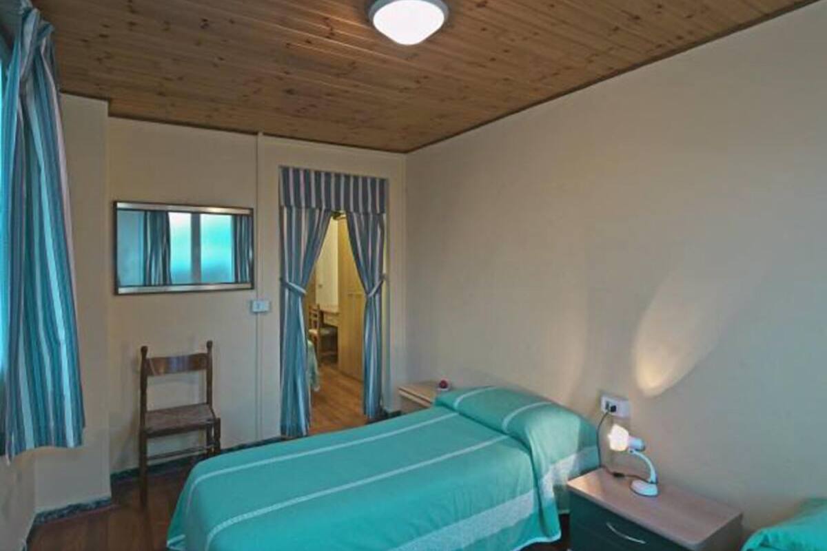 A bedroom with a wooden ceiling, a bed with a green bedspread, a nightstand with a lamp, and a chair.