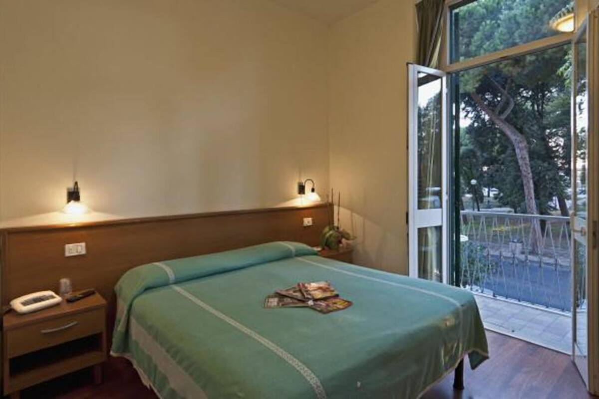 A hotel room with a bed, bedside tables, a window with a view of trees, and a balcony.