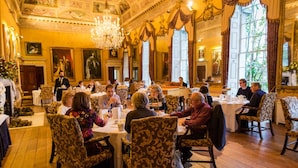 Een formele eetzaal met rijk gedecoreerd interieur, grote ramen en kroonluchters.