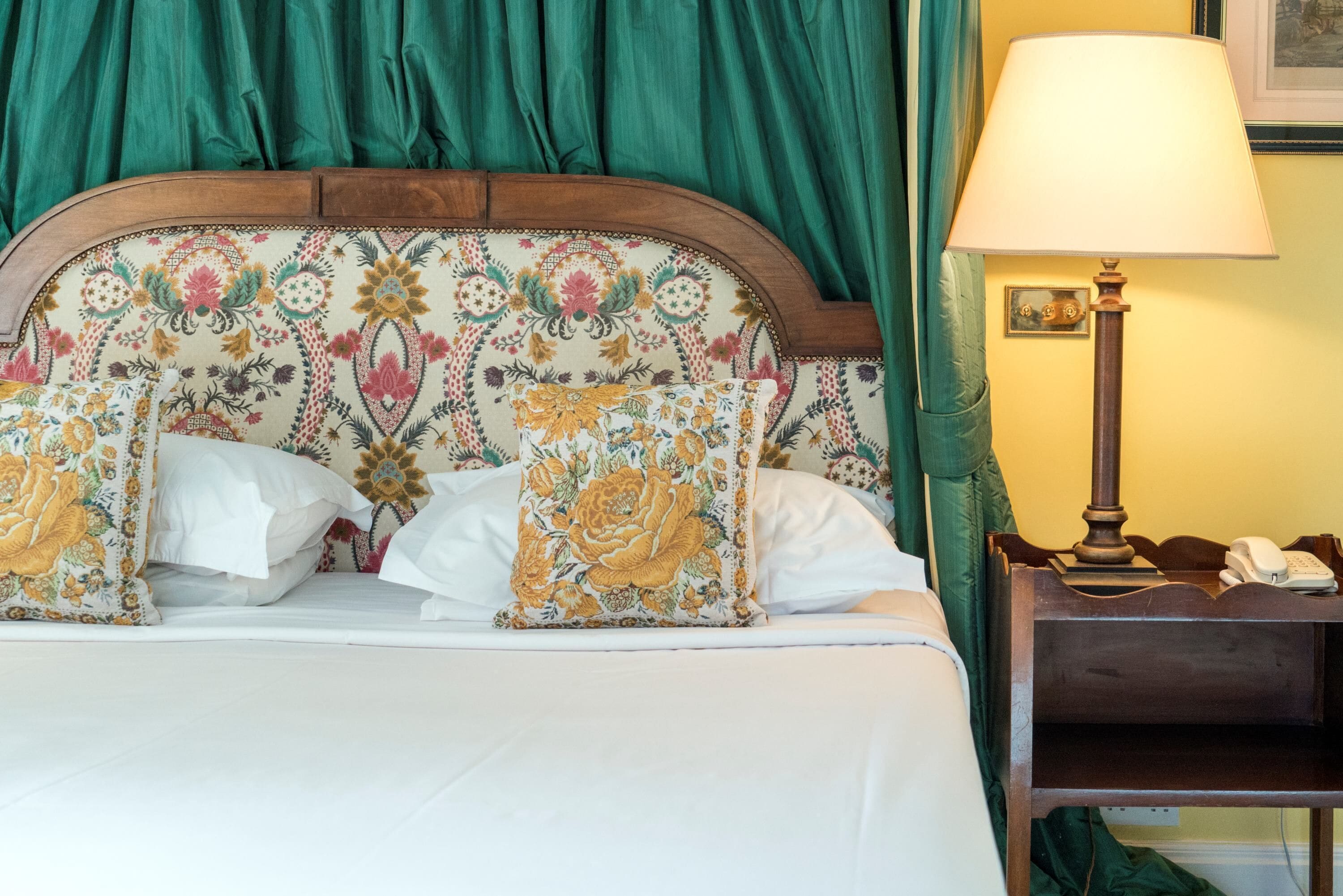 Een bed met een bloemen hoofdbord, witte beddengoed en een nachtkastje met een lamp en telefoon.