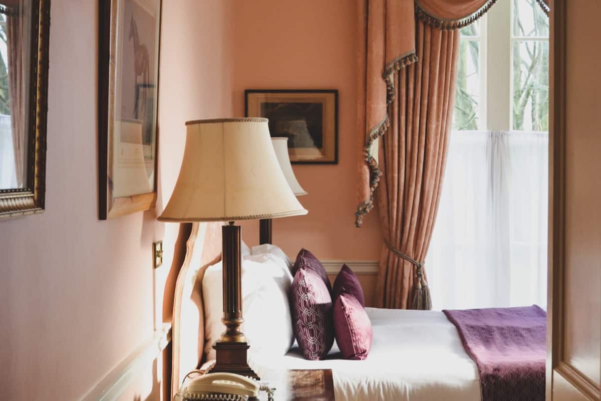 Ein Schlafzimmer mit einem Bett, einem Nachttisch, einer Lampe, einem Fenster mit Vorhängen und gerahmten Bildern an der Wand.