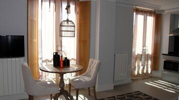 Un coin repas avec une table ronde et deux chaises, un téléviseur fixé au mur et un espace cuisine visible à travers une porte ouverte.