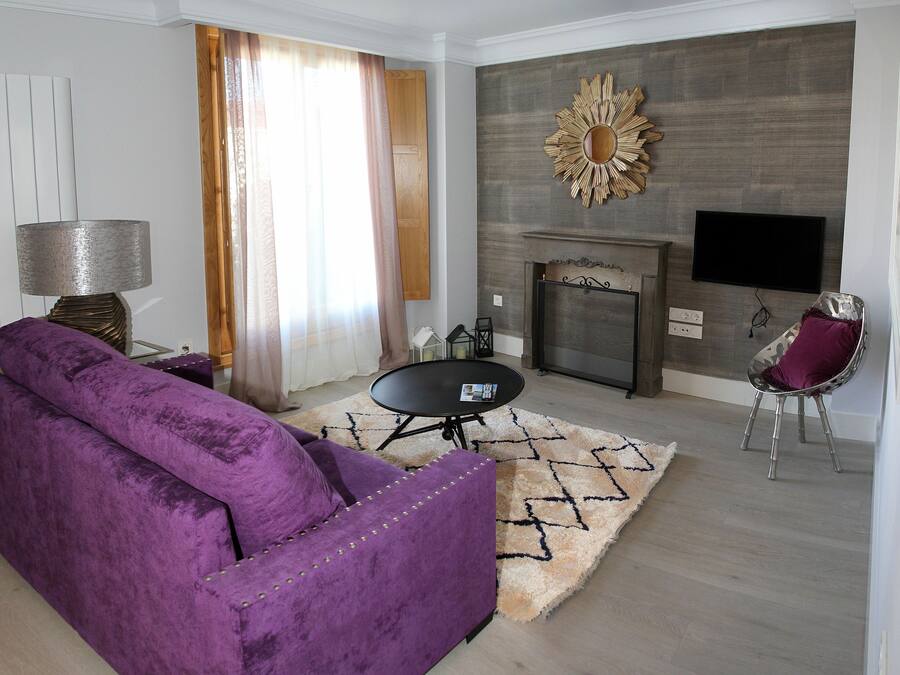 Una sala de estar con un sofá morado, una mesita redonda, un televisor montado en la pared y una chimenea.