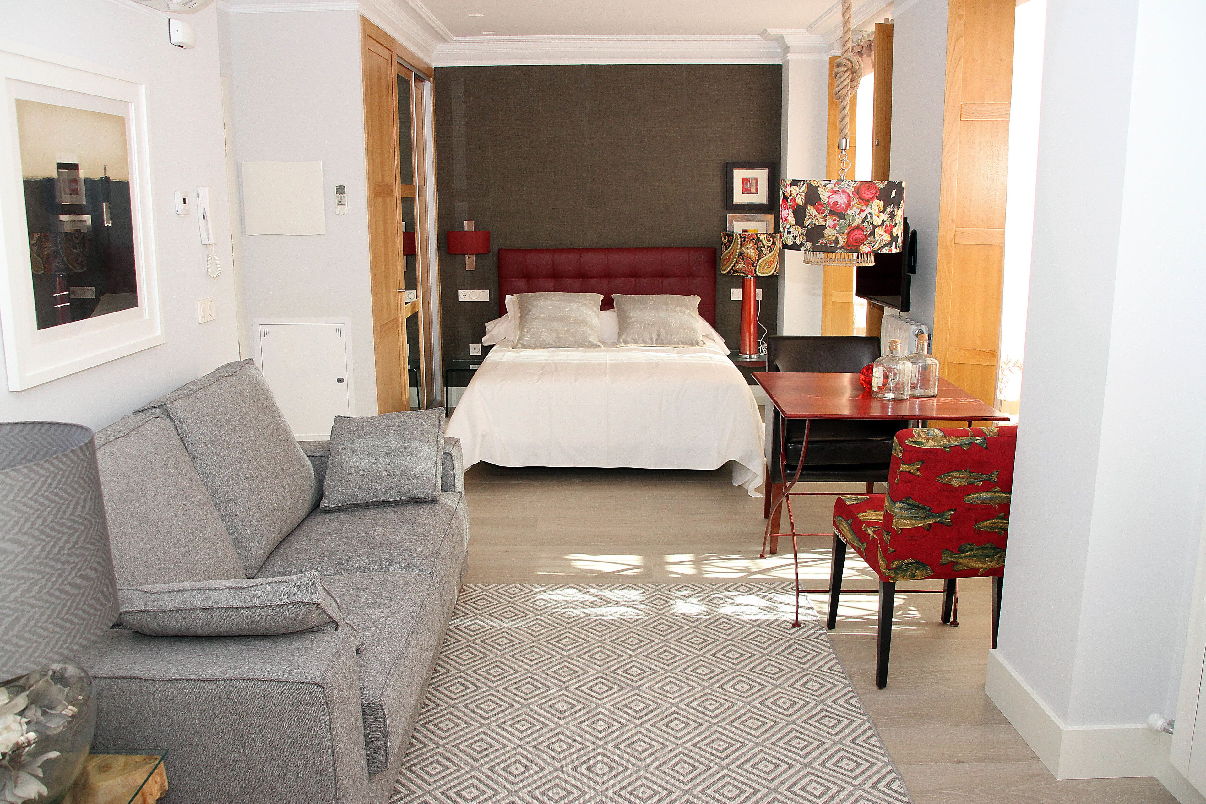 A hotel room with a bed, a grey sofa, a red chair, and a small table with a lamp.