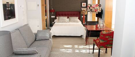 A hotel room with a bed, a grey sofa, a red chair, and a small table with a lamp.