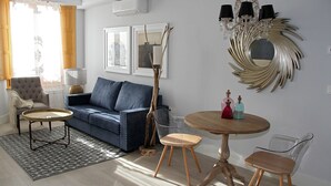 A living room with a blue sofa, a round wooden table, and two chairs. There is a chandelier on the wall and a window with curtains. - Principia Suites (León)