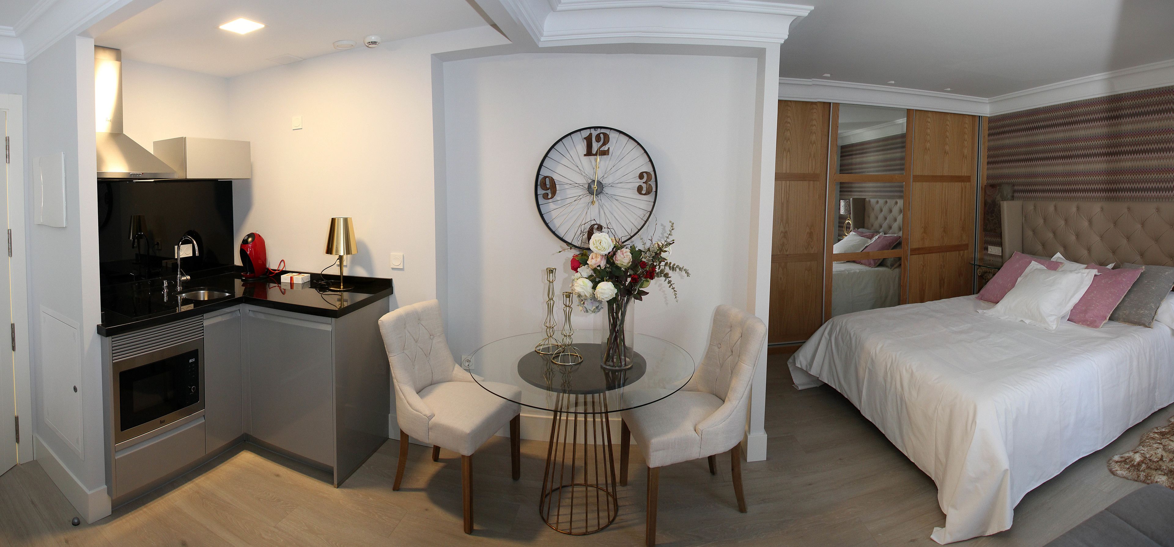 Quarto de hotel moderno com cozinha compacta, área de jantar com mesa de vidro e uma cama grande com cabeceira.